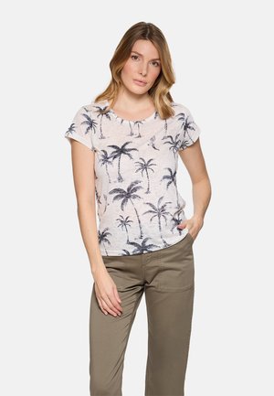 Mujer de pie con una mano en el bolsillo, vistiendo una camiseta blanca con estampado de palmeras negras y pantalones verde oliva sobre un fondo blanco.