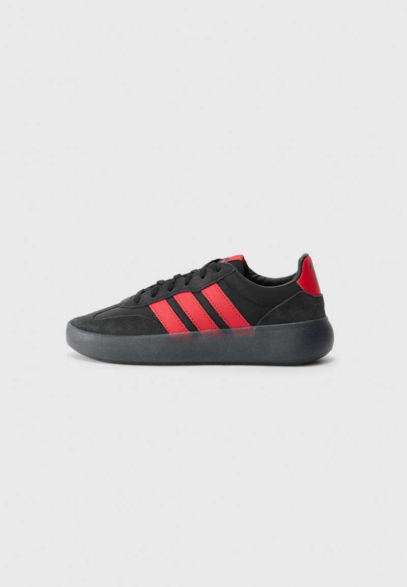 Black and red athletic shoe with a suede and synthetic upper, featuring three prominent red stripes, a rounded toe, and a transparent rubber sole.