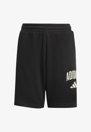Nastolatek w czarnej bluzie z kapturem Adidas, czarnych spodenkach Adidas, w ręku trzyma czarną czapkę Adidas, ma na sobie czarne skarpetki z białymi paskami oraz czarne buty sportowe.