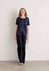 Top corto a maniche corte in satin blu navy abbinato a pantaloni a gamba larga in denim scuro. Il modello tiene una borsa di colore chiaro, in piedi su un pavimento neutro.