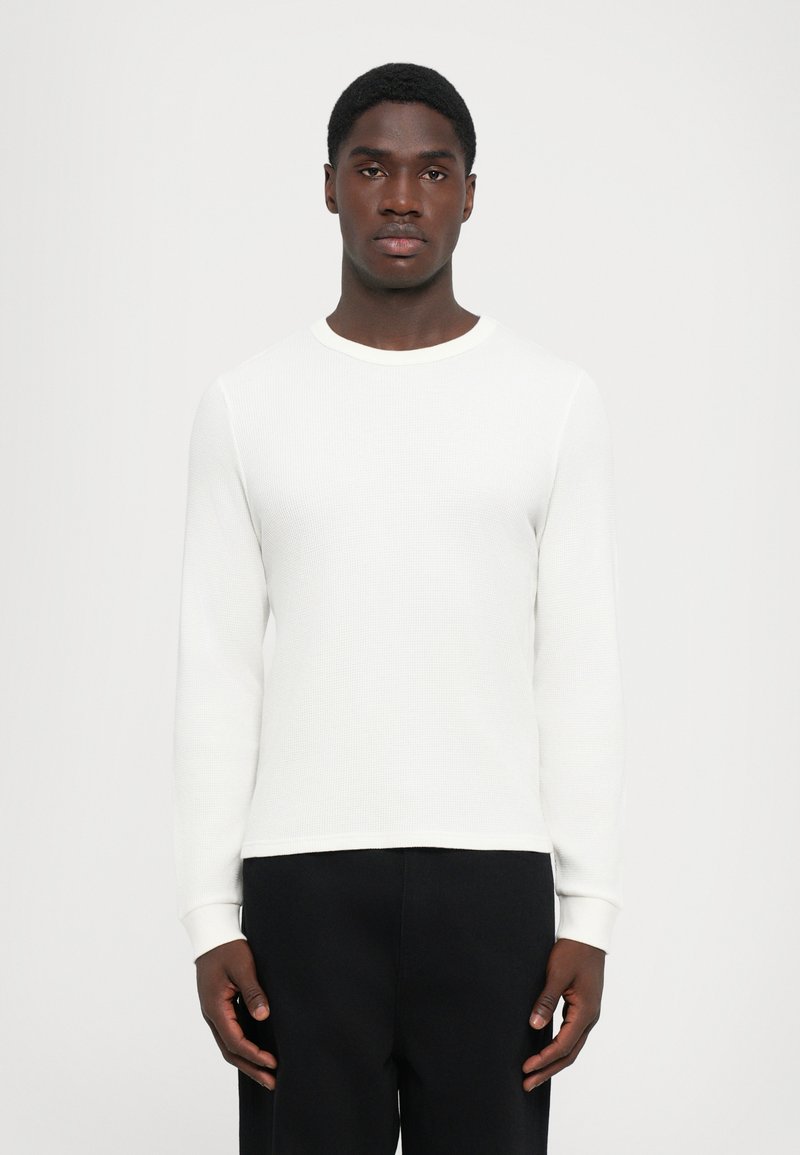 Chemise en tricot côtelé blanche à manches longues, avec un col rond et des poignets ajustés, associée à un pantalon noir. Le tissu a un aspect texturé et léger.
