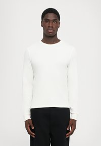 Cămașă albă cu mâneci lungi, cu o textură canelată, cu gât rotund și manșete ajustate, asortată cu pantaloni închiși la culoare. Design simplu, minimalist.