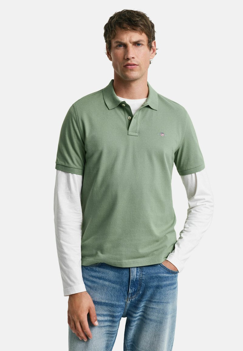 Polo de algodón verde con mangas cortas, que presenta un cuello y una tapeta de tres botones, superpuesto sobre una camisa blanca de manga larga.