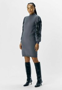 Robe sweat gris à manches longues avec des motifs floraux brodés en noir. Associée à des bottes montantes noires à talons.
