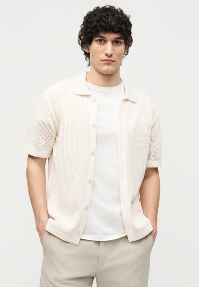 Νέος άνδρας με σγουρά σκούρα μαλλιά που φοράει λευκό, ανοιχτό μάλλινο κοντομάνικο πουκάμισο με κουμπιά πάνω από λευκό μπλουζάκι και μπεζ παντελόνια, με τα χέρια στις τσέπες.