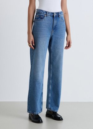 Persona che indossa jeans blu a vita alta con gamba dritta, top bianco, mocassini neri e anelli d'oro su entrambe le mani, in piedi su un pavimento grigio.