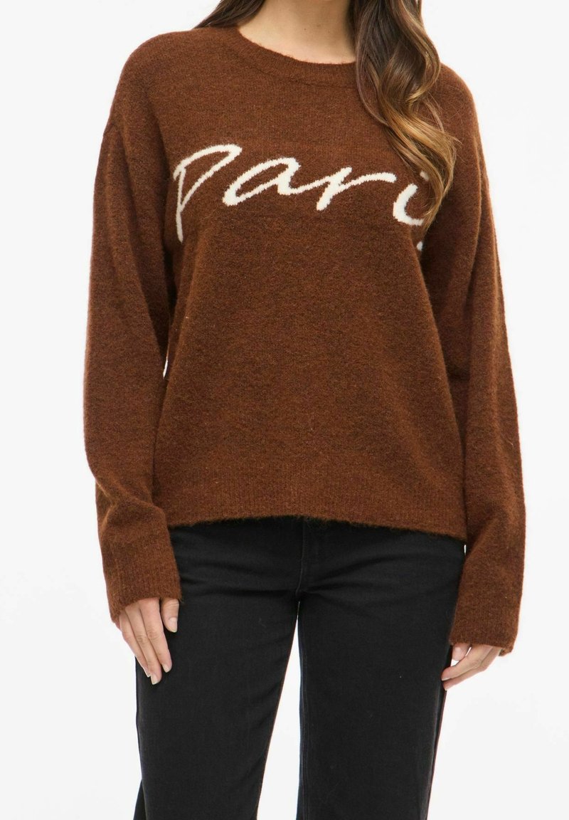 Femme portant un pull marron duveteux avec « Paris » écrit en blanc sur la poitrine et un pantalon noir sur fond blanc uni.