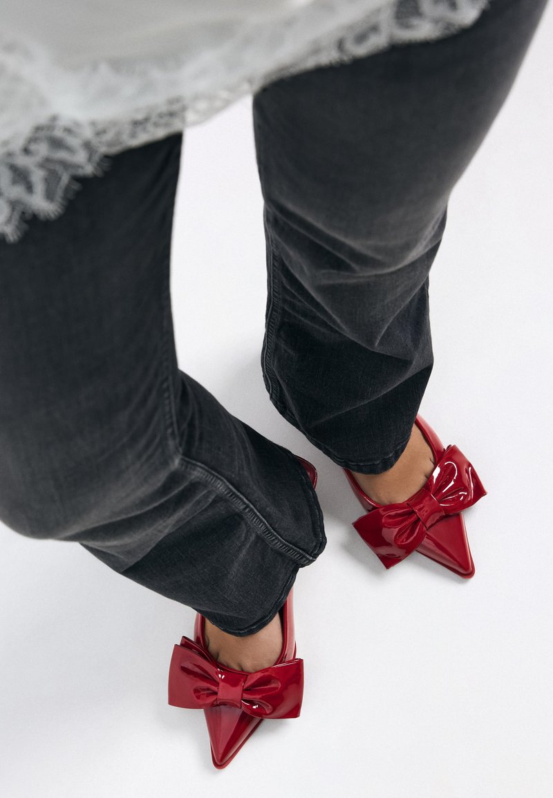 Rote Lacklederschuhe mit spitz zulaufender Spitze und großer Schleife vorne, kombiniert mit dunkelgrauen Schlagjeans und darüber getragenen weißen Lagenstoffen.