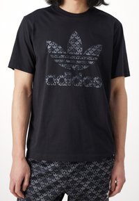 Černé bavlněné tričko s texturovaným černým logem Adidas trefoil a názvem značky ve stejném vzoru. Kulatý výstřih a krátké rukávy.