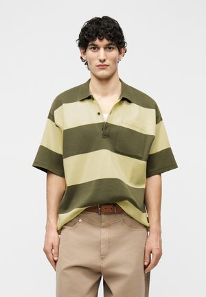 Mladý muž s kučeravými vlasmi, oblečený v zeleno-béžovej polo košeli s širokými pruhmi a béžových nohaviciach, stojaci pred jednofarebným pozadím.