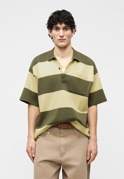 Jeune homme aux cheveux bouclés portant un polo à larges rayures vertes et beiges et un pantalon beige, debout devant un fond uni.