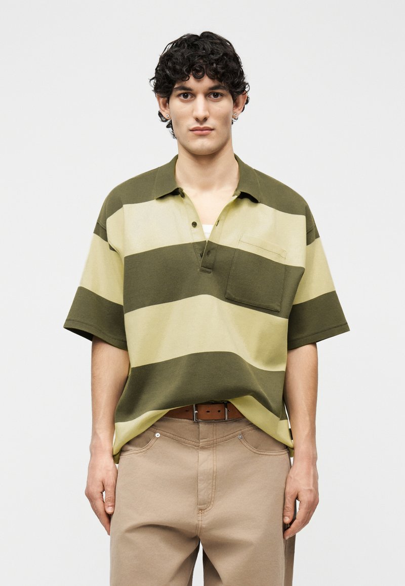 Νέος άνδρας με σγουρά μαλλιά φοράει ένα πράσινο και μπεζ πόλο με φαρδιές ρίγες και μπεζ παντελόνι, στέκεται μπροστά σε μονόχρωμο φόντο.