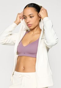 Λευκό φούτερ με φερμουάρ και μακριά μανίκια, κατασκευασμένο από ελαφρύ ύφασμα και με κουκούλα που δένει με κορδόνι. Η εσωτερική στ ρώση αποτελείται από λιλά αθλητικό σουτιέν.