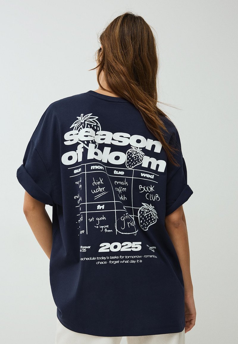 Žena s dlouhými vlasy nosí tmavě modré volné tričko s bílým nápisem "season of bloom", jahodami a týdenním rozvrhem na rok 2025 na zádech.