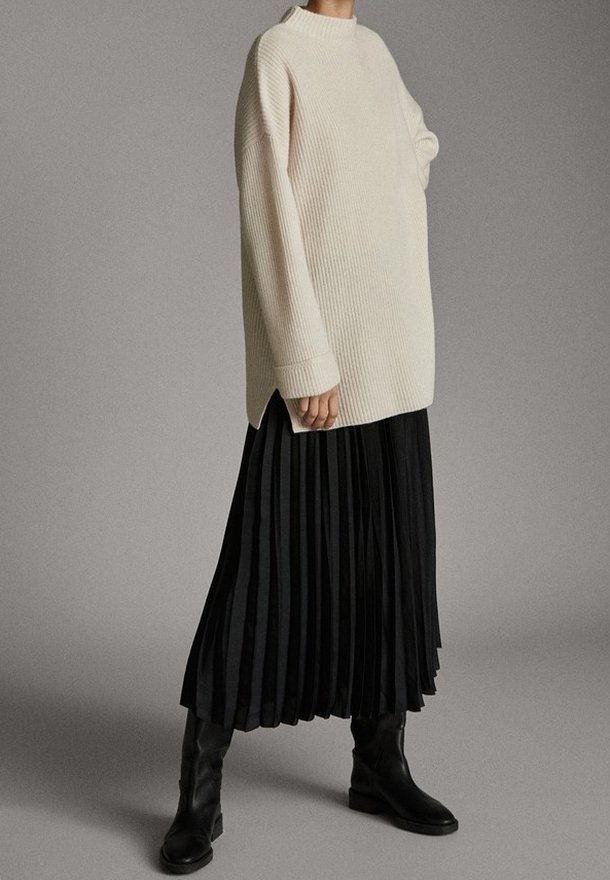 Personne portant un pull côtelé crème oversize, une jupe midi plissée noire et des bottines en cuir noir, debout devant un fond gris uni.