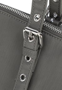 Grå tygväska med en texturerad finish, prydd med silverfärgad hårdvara, en dragkedja och nitar för remfästen.