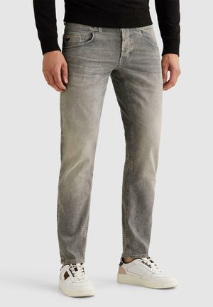 Homme portant un jean slim gris délavé, un haut à manches longues noir et des baskets blanches, debout devant un fond uni.