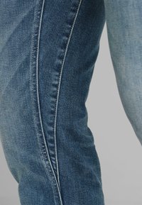 Close-up of a person's leg wearing blue denim jeans with visible stitching and fabric texture against a plain gray background.