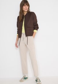 Femme portant une veste zippée marron par-dessus un pull vert clair, un pantalon beige avec cordon de serrage, des chaussettes blanches et des baskets beige avec des touches de vert.