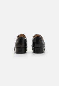 Chaussures habillées en cuir noir avec détails brogue, bout arrondi et petit talon. Texture lisse avec semelles intérieures en cuir marron contrastant et coutures décoratives.