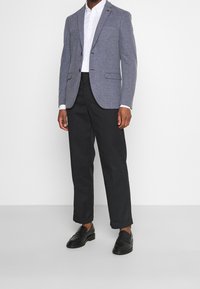 Man som bär en grå kavaj, vit skjorta, svarta byxor med uppvikningar, och svarta loafers, står mot en enkel vit bakgrund.