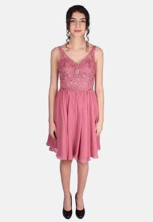 Jonge vrouw met krullend haar die een mouwloze roze jurk draagt met kanten en paillettendetails, staand en zwarte pumps dragend.