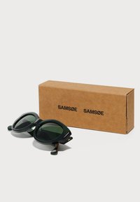 Green, oversized sunglasses with rounded lenses and contrasting black accents, placed next to a brown cardboard box featuring "SAMSØE."