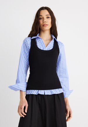 Kobieta z długimi kręconymi włosami, ubrana w niebieską koszulę w paski, na którą nałożona jest dopasowana czarna kamizelka bez rękawów oraz czarne spodnie, na białym tle.