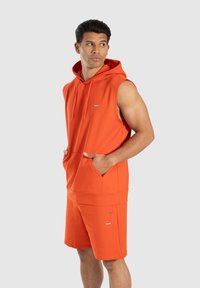 Uomo che indossa un hoodie arancione senza maniche e pantaloni corti abbinati, in piedi con le mani in tasca e che guarda di lato contro uno sfondo neutro.