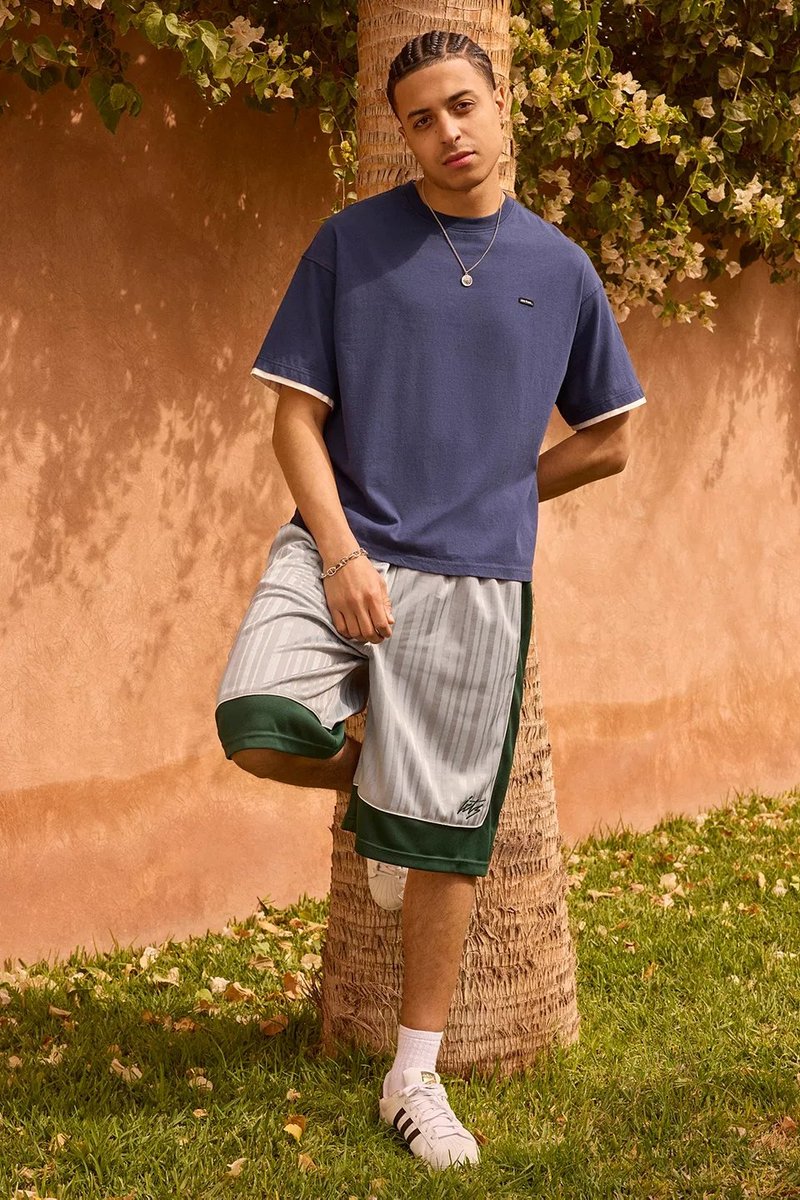 Marineblaues T-Shirt mit kurzen Ärmeln und weißen Akzenten, kombiniert mit grauen gestreiften Shorts mit grünen Details. Das Model trägt weiße Sneakers.