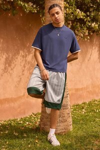 Marineblåt kortærmet t-shirt med hvide detaljer, kombineret med gråstribede shorts med grønne detaljer. Modellen har hvide sneakers på.