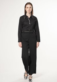 Camicia nera a bottone con rifiniture argentate, abbinata a pantaloni neri a gamba larga. Scarpe con cinturino argentato completano l'outfit. Sfondo semplice.