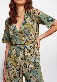 Jumpsuit floral en verde, con patrones azules y dorados, mangas cortas, un profundo escote en V y un detalle de cintura atada. Tejido texturizado.