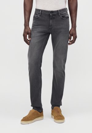Homme portant un jean gris foncé et des chaussures marron à enfiler, debout devant un fond clair uni.