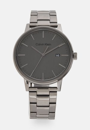 Montre Calvin Klein avec un boîtier et un cadran en métal gris, des index horaires minimalistes, et un bracelet en acier inoxydable argenté. Affichage de la date à 3 heures.