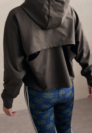 Person iført en mørk cropped hættetrøje med udskæring på ryggen og blå leggings med gult palmetræsmønster og hvide sidelængder.