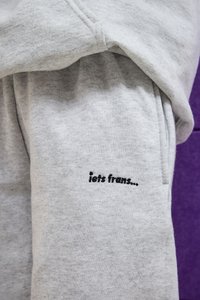Hellgrauer Sweatpant-Bereich mit schwarzem besticktem Text "iets frans..." auf dem Stoff, vor einem lila Hintergrund.