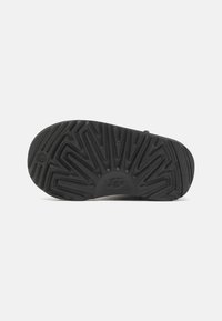 Semelle en caoutchouc noir avec un motif en zigzag texturé et le logo UGG au centre. Conçue pour offrir adhérence et durabilité.