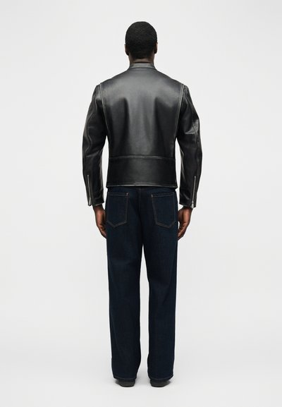 Άνδρας που φοράει μαύρο δερμάτινο μπουφάν και σκούρο μπλε τζιν, στέκεται γυρισμένος με την πλάτη προς ένα απλό φωτεινό φόντο.