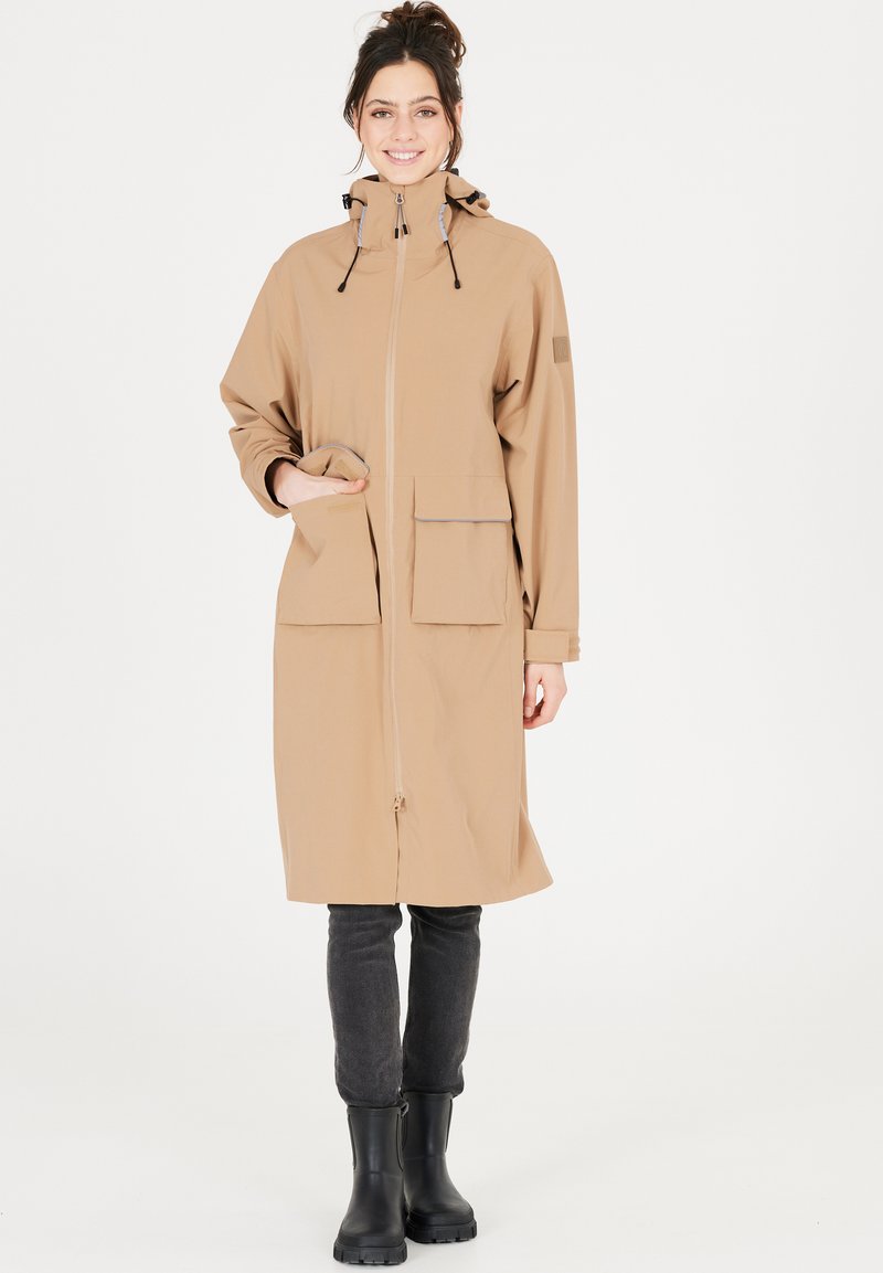 Beige vandtæt jakke med hætte, frontlynlås, to store lommer og lange ærmer. Båret over mørke jeans og sorte gummistøvler.