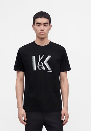 Mladý muž v čiernom tričku s veľkými striebornými "IK" písmenami a štýlovou profilovou grafikou na jednofarebnom bielom pozadí.