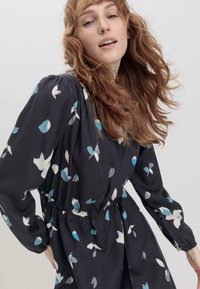 Blumenkleid aus dunklem Stoff, mit blauen und weißen Blattmustern, langen Ärmeln und gerafftem Taillenbereich für eine entspannte Silhouette.