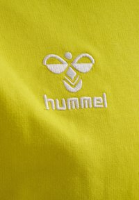 Hvid broderet bi-logo og "hummel" tekst på en lys gul stofbaggrund.