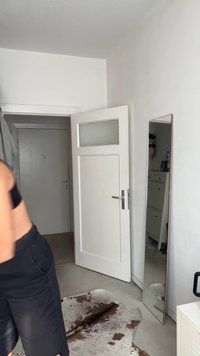 Porta branca com painel de vidro fosco, espelho de corpo inteiro com moldura de madeira, paredes claras e um tapete castanho texturizado num chão neutro.