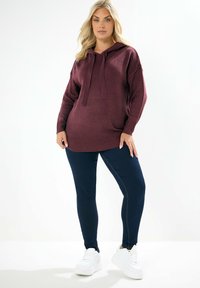 Burgundy hoodie med en strukturerad stickning, långa ärmar och en framficka, parat med mörka leggings och vita sneakers.