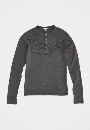 Donkergrijs henley-shirt met lange mouwen, vier knopen en grote cursieve tekst "Blessed" over de borst.