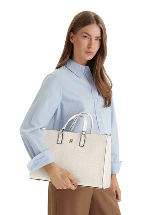 Donna con camicia azzurra che tiene una grande borsa bianca testurizzata con doppi manici su uno sfondo bianco uniforme.