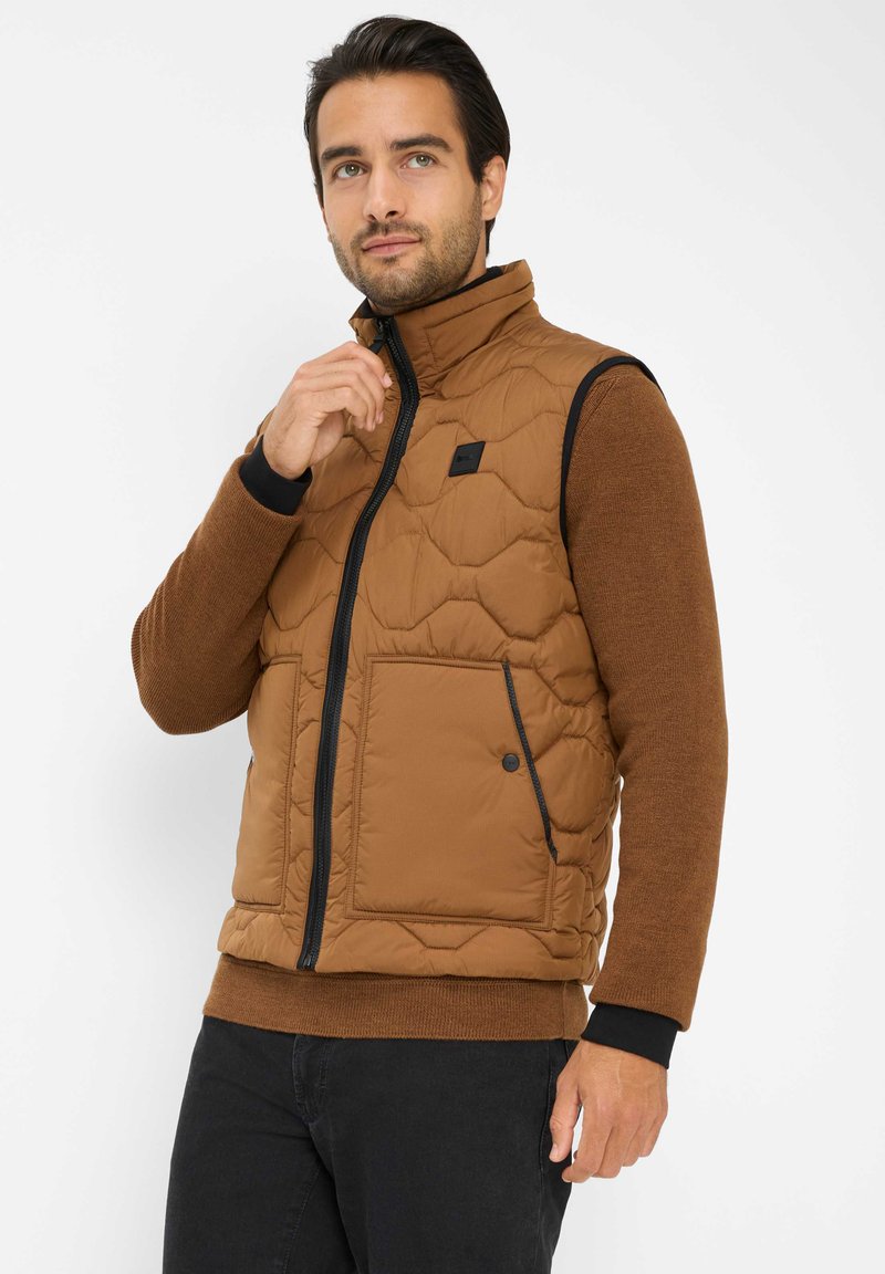 Gilet imbottito marrone con colletto alto, zip nera e tasca frontale. Indossato con un maglione marrone, caratterizzato da polsini e orlo a coste. Pantaloni scuri.