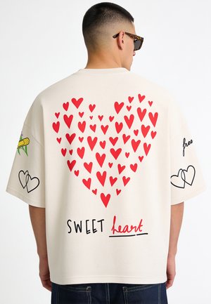 Homme portant une chemise blanche oversize avec un motif de cœur rouge formant un grand cœur et le texte "SWEET heart" dans le dos.