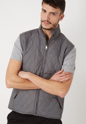 Chaleco gris acolchado con cuello alto, cremallera frontal y dos bolsillos laterales. Camiseta gris de manga corta debajo. Textura suave.